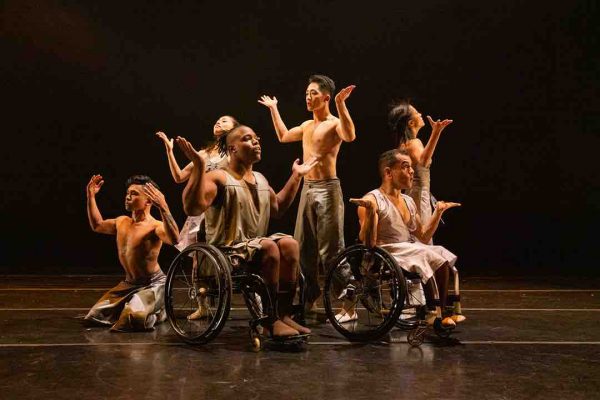 Sixe disabled and non disabled dancers reach their hands outwards with bent elbows in a clump; they wear silver, flowing costumes and perform on a black marley stage.