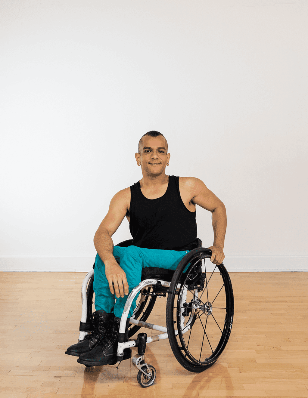 JanpiStar is a Latinx wheelchair user with light brown skin, long arms and a beautiful smile. They are wearing a black tank top, blue turquoise pants and black boots.