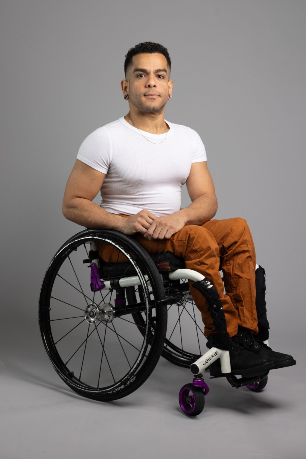 2025AXIS_RobSuguitan_Janpi_Portrait JanpiStar poses for a full-body portrait. They are a Latinx wheelchair user with light brown skin and long, muscular arms. They are wearing a tight white t-shirt and brown pants.