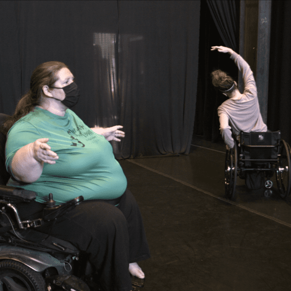 Heidi cash sits in her power wheelchair, reaching two hands outwards. Ashley tilts to the side in her manual wheelchair, lifting one arm up into a curve.