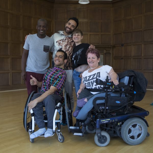 A diverse group of five disabled and non-disabled dancers and choreographers pose in a studio together. The studio has wooden floors and walls; a blue wig lies on the floor in the background.