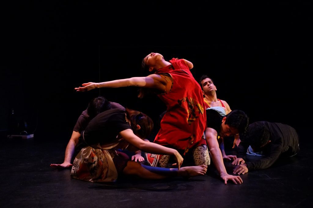  AXIS performs a work in progress by Neve Mazique-Bianco from the 2019 Choreo-Lab. Lani Dickinson wears a red dress and reaches outwards with one arm while kneeling on the ground. Other dancers lie on the floor beside her, heads towards the ground, with the exception of JanpiStar who mirrors her expression in the background.