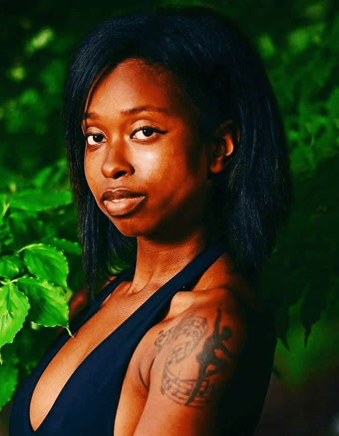 Ellice Patterson poses for the camera, surrounded by green leaves. Elise is a black woman with straight black hair and a tattoo of a dancer surrounded by music notes on her shoulder. She is wearing a black halter top.