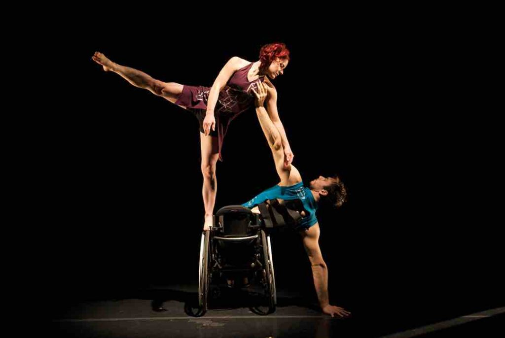 Joel, a dancer who uses a wheelchair, places one hand on the floor in a side lunge while bringing his other arm to support Sonsherée. Sonsherée balances one leg on Joel's chair while extending her other leg; she looks towards Joel as she holds the length of his arm for support. Joel wears a sleeveless turquoise shirt; Sonsherée wears a burgundy jumpsuit. The dancers perform in bright stage lighting juxtaposed to a dark background.