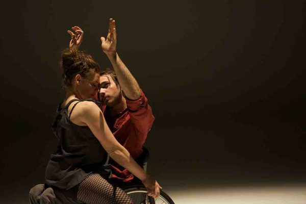 oel sits in his wheelchair and lifts his arms while looks up; Tanja hits on his lap and closes her eyes and smiles as tilts her head forward. Joel wears a maroon dress shirt, Tanja wears a loose gray top. The dancers perform in dramatic studio lighting.