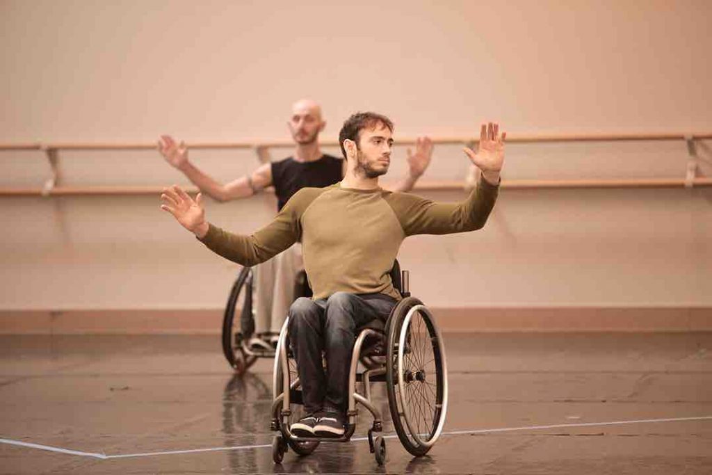 Two dancers using wheelchairs bring two arms upwards with bent elbows and look to the right, wearing street clothes in a dance studio. Joel is in the foreground of the image, while Marc dances in the background.