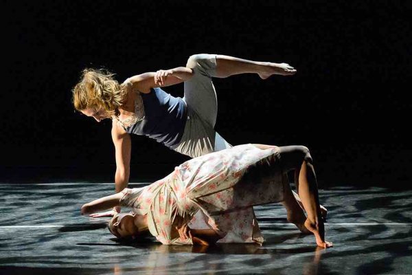 Julie pushes her torso upwards with her legs as she rolls towards the right on the floor. Sophie balances in a side plank above her; she lifts one and and one arm up towards Julie. Sophie wears a camisole with a lace pattern, Julie wears a floral dress as they dance in dappled theater lighting.