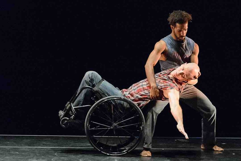 Nick holds a plié in second position as Dwayne leans back in his wheelchair to rest his back on Nick's upper thigh. Dwayne reaches out with one hand towards the audience. Both dancers wear jeans and plaid sleeveless shirts.