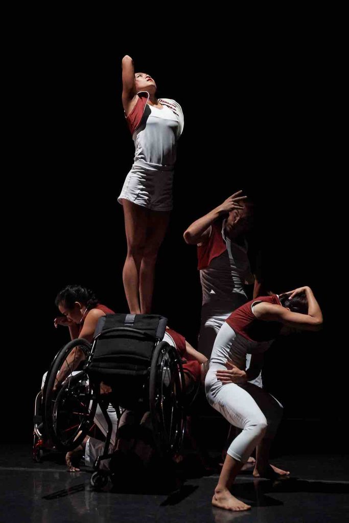 Five disabled and non-disabled dancers sit / stand in a clump with chests contracted inwards as they raise their hands to lowered faces. Dwayne tilts forward in his wheelchair and Lani stands on his back she raises her upper limb into the air. All Five Dancers wear white costumes with maroon vests.