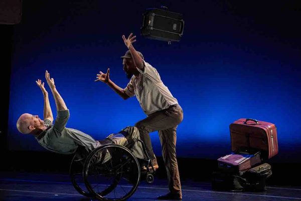 Dwayne leans back in his wheelchair with two arms raised in front of his head; James holds one foot on the footrests of Dwayne's chair to balance, while he places his hands in a similar position. Suitcases hang from the ceiling and are placed behind them.