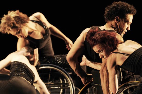 In the foreground, Sonsherée Giles rests her head on Rodney Bell's shoulder, who is turning forward in his manual wheelchair. In the background, Janet Das rests her head in the arm of Alice Sheppard, who leans to the side in her manual wheelchair. The dancers wear black costumes with sequins accents.