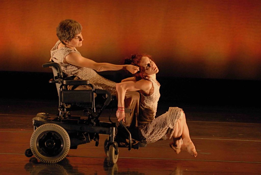 Sonsherée sits on the feet of Bonnie's power wheelchair with her feet raised; Bonnie reaches to touch the side of Sonsherée's head in a tender gesture. The dancers wear beige costumes that have geometric patterns.