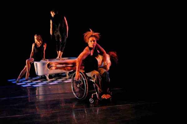 Alice is photographed mid-turn in her wheelchair; Sonsherée balances on her lap in a sideways plank. In the background Juliana and Sebastian pose seductively on a white claw-footed bathtub a white and black checkered floor.