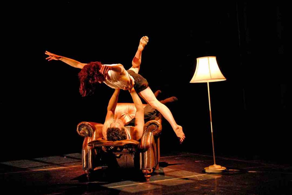 Rodney lies on a brown leather easy chair and lifts Sonsherée upwards; Sonsherée extends all four limbs out with out leg bent and reaching up. There is a lamp to the right of the dancers.