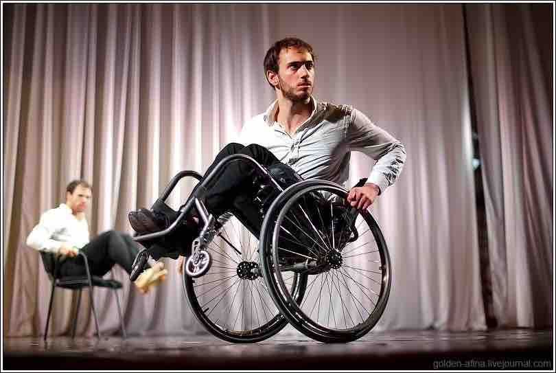 In the foregound, Joel balances a wheel in his wheelchair; in the background, Sebastian translates his movement while sitting in a four-legged chair.