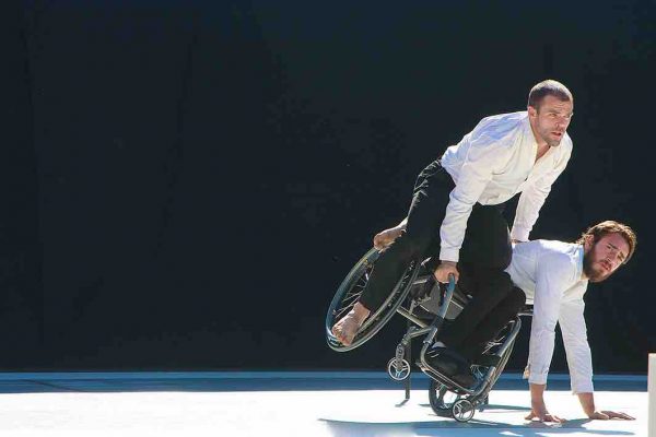 Joel leans into a side tilt in his wheelchair with two palms on the ground and one wheel in the air. Sebastian sits on the side of Joel's chair that is raised in the air as he looks forward. Both dancers wear white collared shirts and black pants.