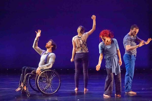 Four disabled and non-disabled dancers perform on a stage with blue lighting and a black marley dance floor. Joel, a wheelchair user, lifts one hand up while the other one rests on his chair. Juliana mirrors his gesture while standing with her back to the camera, Sonsherée looks down towards one hand and Sebastian looks down towards two hands. The dancers wear costumes with various shades of blue and gray.