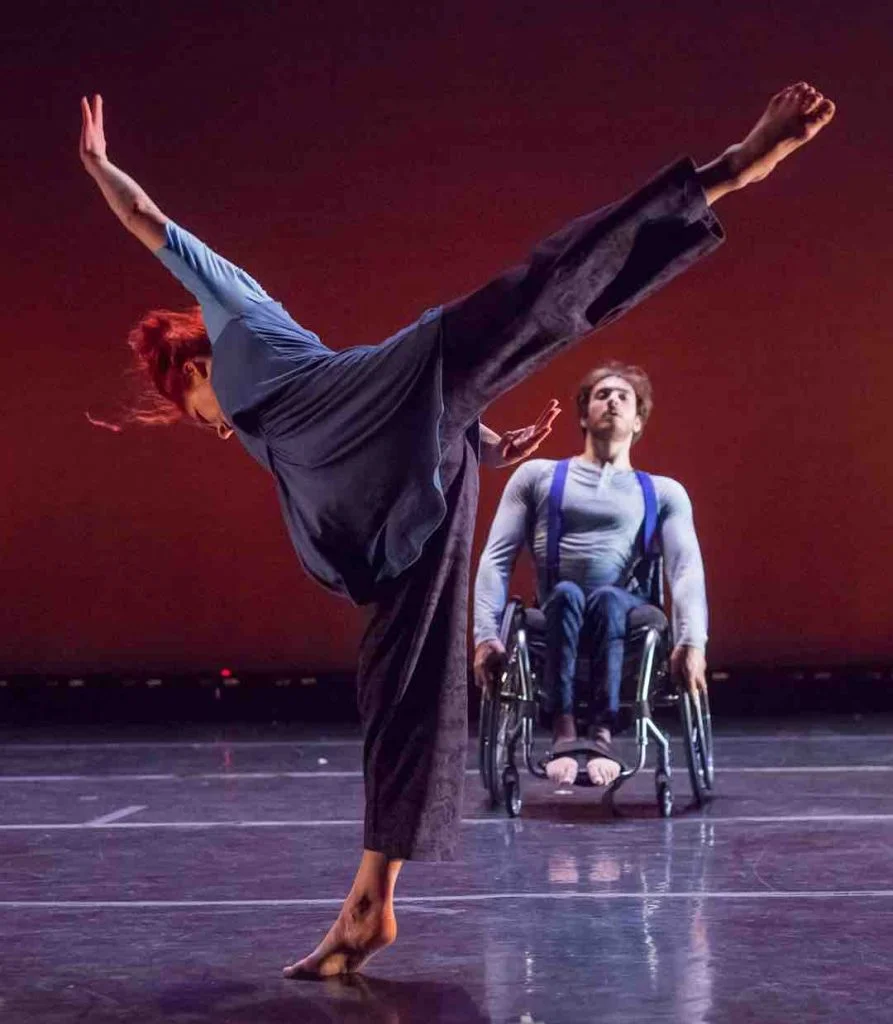 In the front of the image, Sonsherée extends an arabesque in relevé with hands reaching forward. In the background, Joel leans back while sitting in his wheelchair.