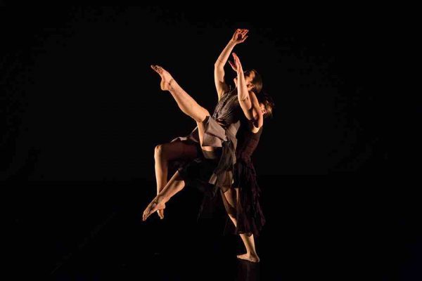 Liv holds Julie in a lift as Liv brings one leg forward and space. Julie gestures upwards with both upper limbs, extending her legs forward as well. Both dancers wear textured dresses with brown and maroon coloring.