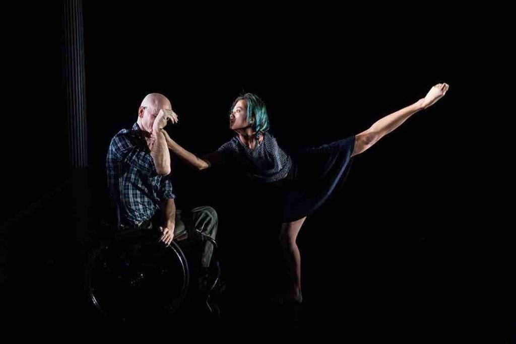 Lani places one hand on the shoulder of Dwayne as she extends one leg back in a high arabesque. Dwayne reaches one arm across his chest to a wheel of his wheelchair, and brings his other one up into a curved wrist gesture in front of his face. Dwayne wears a plaid shirt and olive green pants so my: Lani has turquoise dyed hair and wears a blue skirt and top.