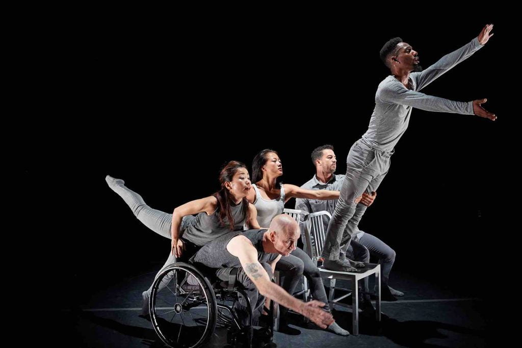 Five disabled and non-disabled dancers lean forward together in a clump. Yuko extends one leg back as she places her torso on Dwayne's back, James tilts forward while standing on a chair as Lani holds his leg for balance. The dancers wear simple gray costumes.