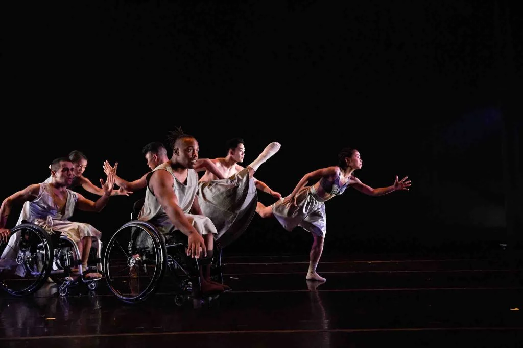 Five disabled and non-disabled dancers lean forward together in a line with outstretched hands. Standing dancers balance on one foot and extend their other leg as they move. The dancers wear silver, flowing costumes.