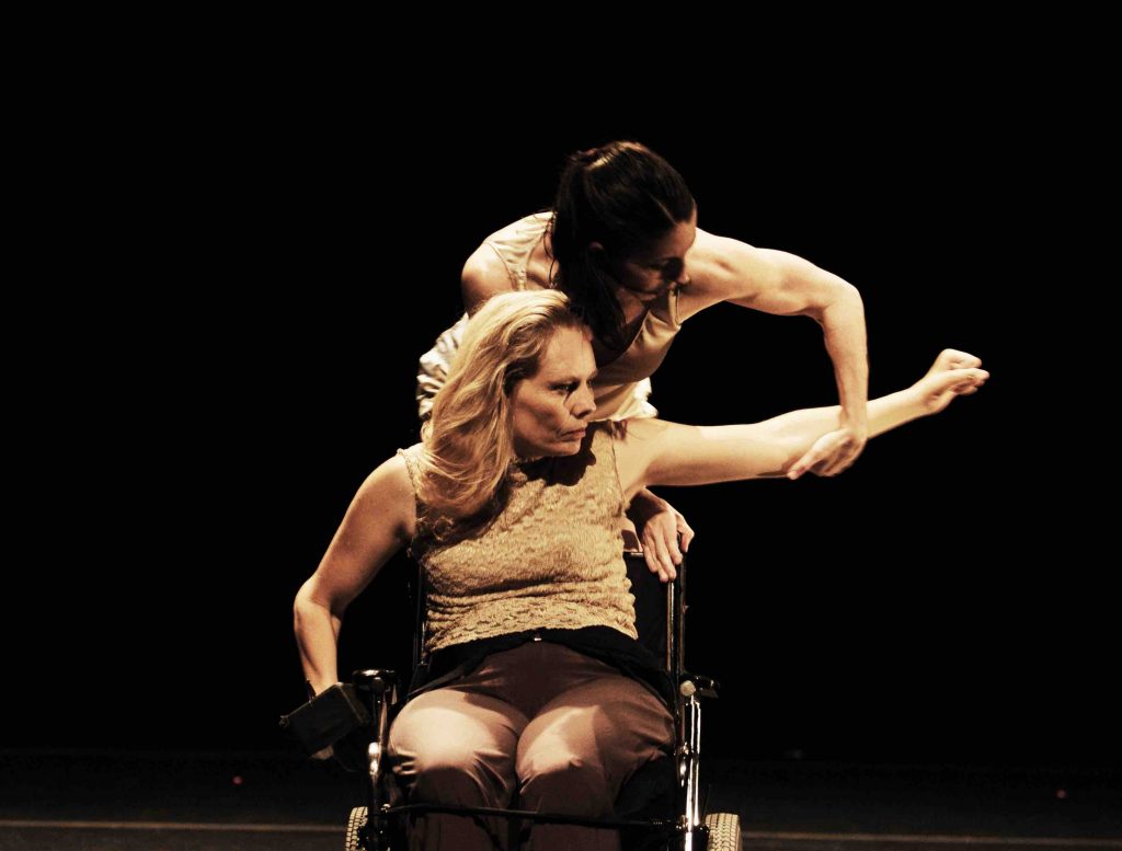 Judith reaches her arm to the side with a fierce expression on her face, she is sitting in a manual wheelchair. Janet stands behind her and holds her elbow.