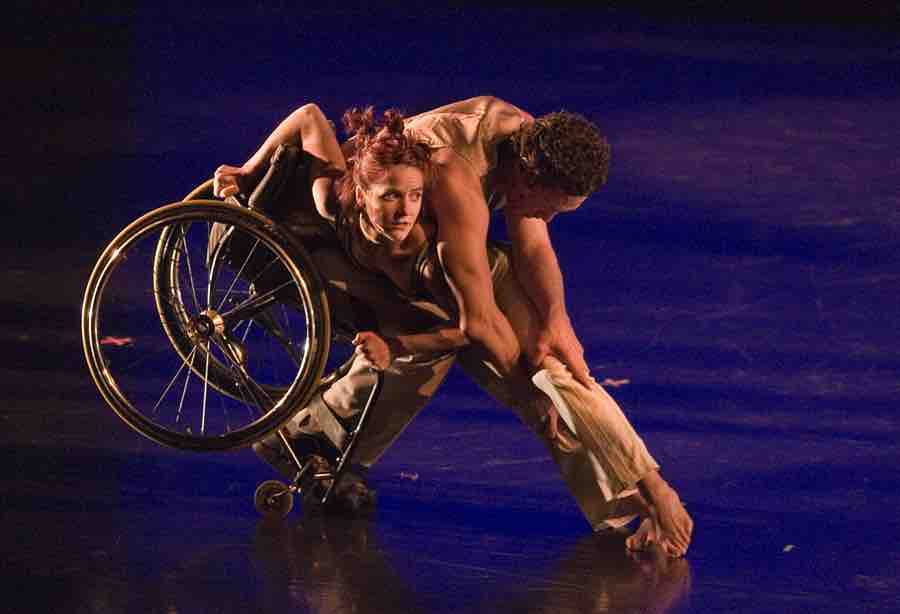 Rodney balances in a forward tilt in his wheelchair; he holds on to the legs of Sonsherée who crouches on his lap with her torso and holds her feet on the floor in line with his body.
