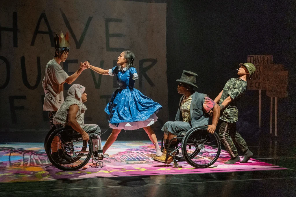 Five disabled and non-disabled dancers are dressed like characters from Alice in Wonderland set in modern day San Francisco. Lani balances on the footrests of DeMarco and Janpi, she holds the hand of Bradford for support as AG leans backwards behind DeMarco. There is a psychedelic palm tree illustration on the ground and a sign in the background reading "Have You Ever Felt."