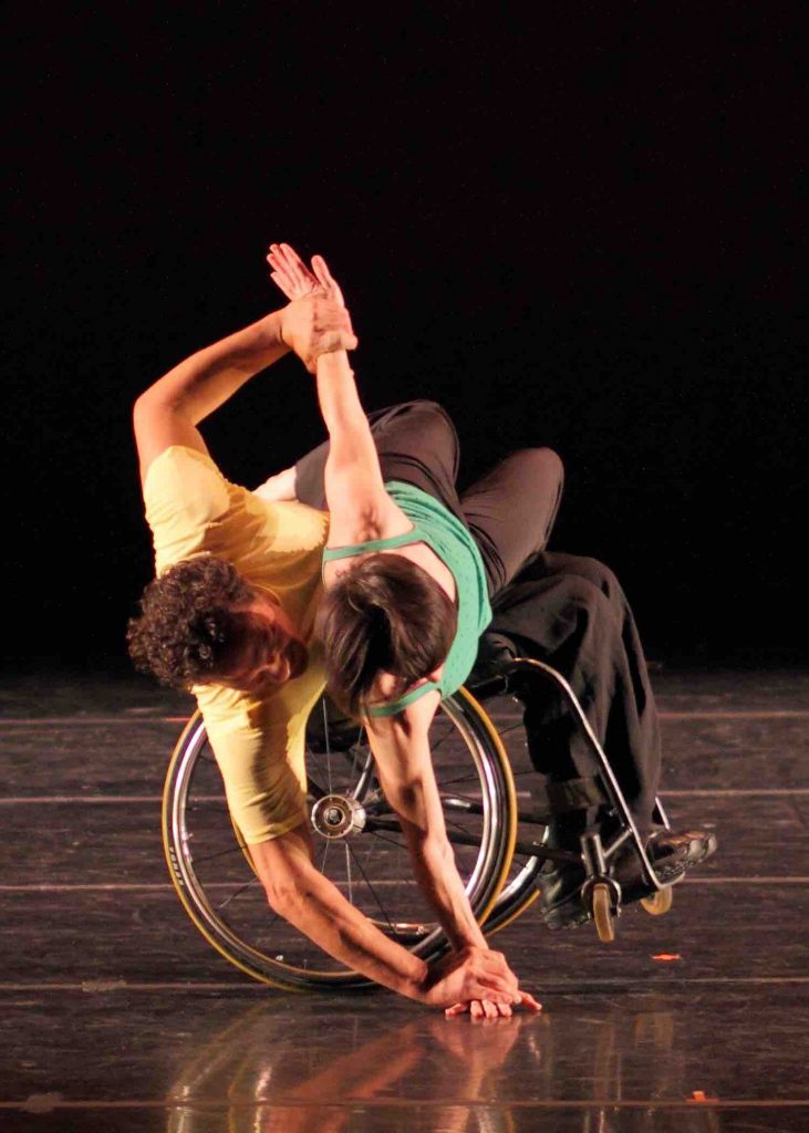 Rodney balances in a wheelie in his wheelchair while placing on hand on the ground to his side. Janet lies across his lap and places one hand next to his on the ground, while extending her other hand upwards to meet Rodney's grip. The dancers wear yellow and green shirts and black pants.