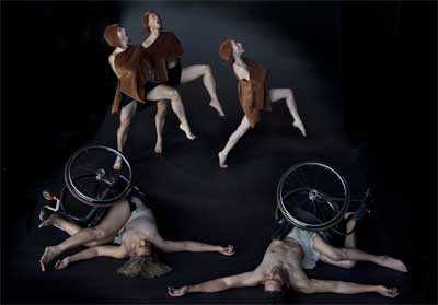 In the background, three non-disabled dancers lunge forward into space with one bent knee; they wear light brown cloths over their torsos with matching hats. In the foreground, Alice Sheppard and Rodney Bell lie on their backs in their manual wheelchairs, their torsos shifted to opposing sides.