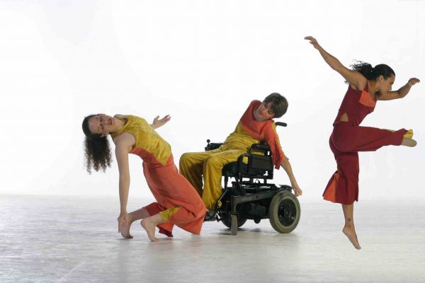 Alisa holds a backwards tilt as she reaches to the floor with on arm, Bonnie reaches sideways in her power wheelchair, Stephanie leaps forward; she has a lower limb difference. All three dancers wear costumes of yellow, red and orange.