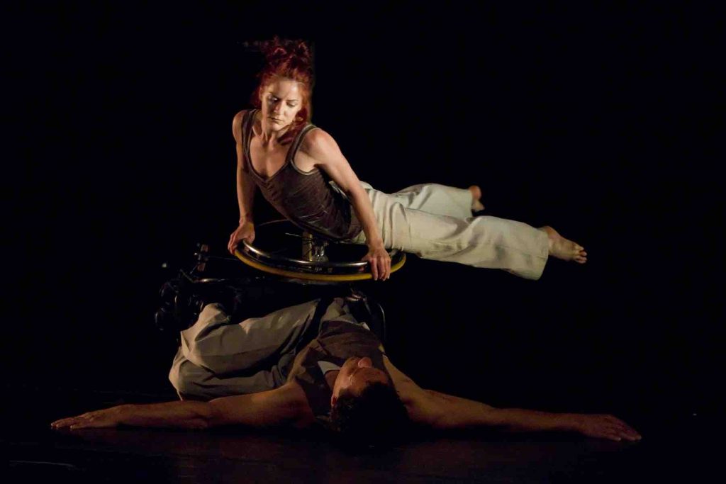 Rodney lays on back side in his wheelchair, with his arms spread out wide and wheelchair/torso turned to the side. Sonsherée balances her toros on his right wheel as she spins around it's axis.