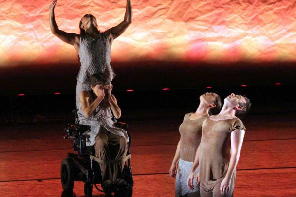 Bonnie brings her hands towards her face, while Stephanie stands with her arms outstretched to the sky on top of Bonnie's power wheelchair. Sean & Renee kneel perpendicular to the two dancers, raising their chests upwards with arms to the side. The dancers perform on a stage with a textured brown background that is softly illuminated from below.