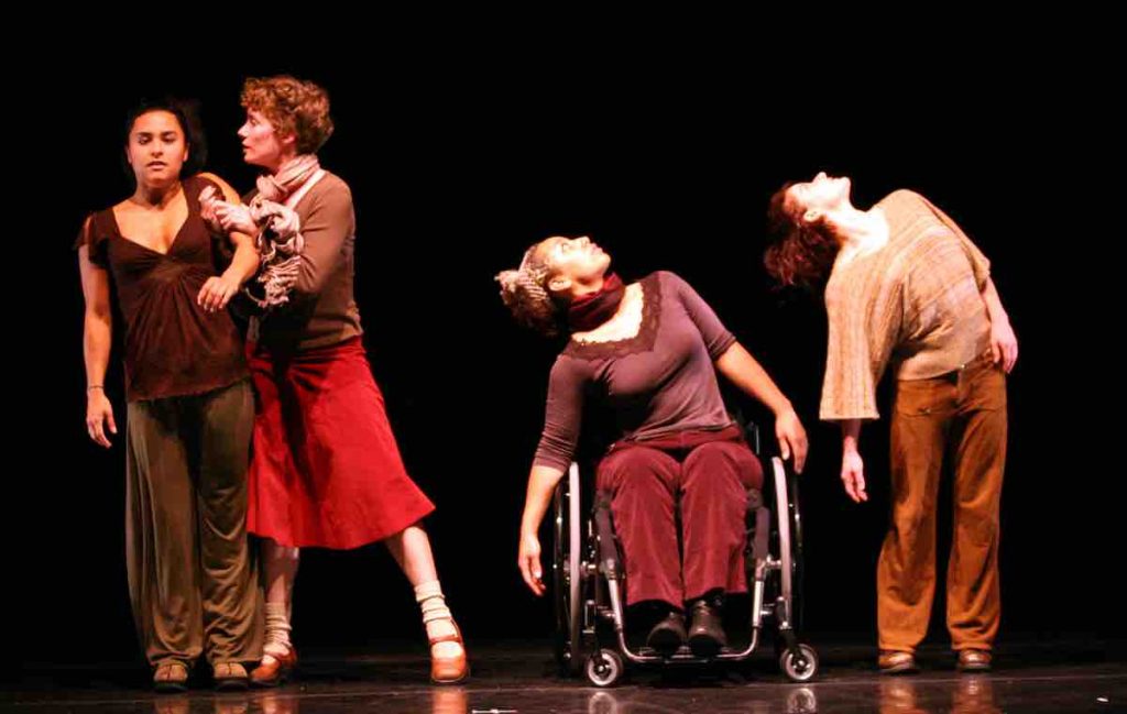 Margaret holds the shoulder of Stephanie, who reaches one arm forwards. Alice and Sonsherée tilted to the side and look up words; Alice in her wheelchair and Sonsherée standing.