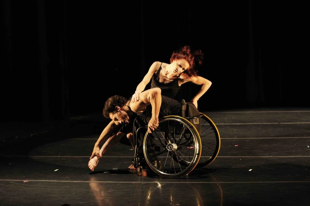 Rodney leans forward in his wheelchair, grabbing the foot of Sonsherée as she leans against him and pivots forward. Both dancers wear all black.