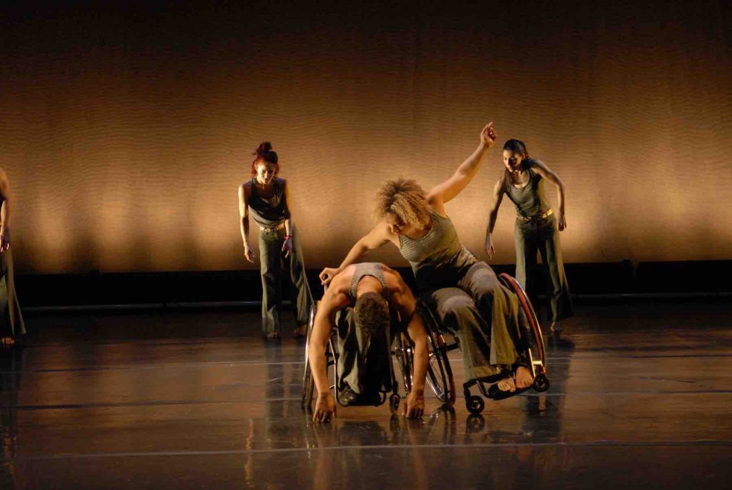 Alice Sheppard does a sideways tilt in her wheelchair, lifting one arm up and holding onto Rodney bell for balance. Rodney leans forward in his wheelchair. In the background, Sonsherée Giles and Janet Collard tilt to the side with arms handing down. Dancers wear olive green costumes.
