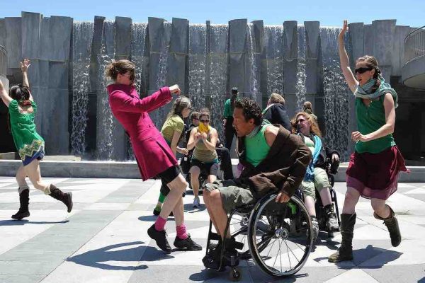 A cast of disabled and non-disabled dancers run and wheel around each other an a concrete outdoor square with a sculptural gray concrete fountain wall behind them. The dancers wear costumes with green, red and yellow hues.