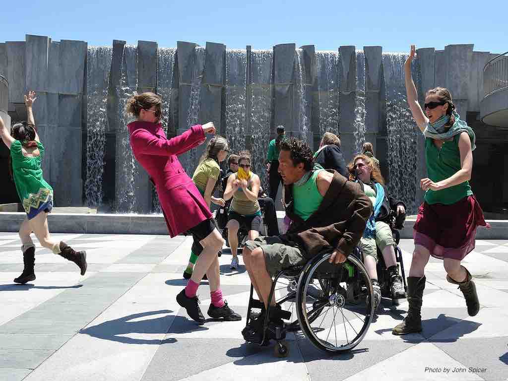 A cast of disabled and non-disabled dancers run and wheel around each other an a concrete outdoor square with a sculptural gray concrete fountain wall behind them. The dancers wear costumes with green, red and yellow hues.