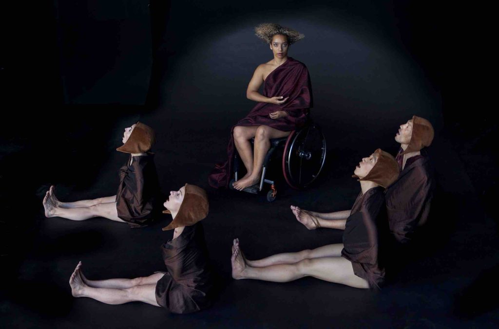 Alice Sheppard Sits in her wheelchair with Palms resting open on her lap; she wears a flowing maroon robe and her hair is arranged into a geometric triangular shape. In front of her, foyr dancers sit with legs out and heads raised with open mouths; they are wearing brown cloths that cover their arms and light brown caps.