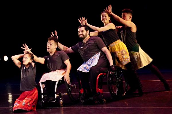 Five disabled and non-disabled dancers sit and stand in a vertical line; they place hands over each other's heads and make silly faces. The dancers wear black AXIS t-shirts and multi-colored earrings.