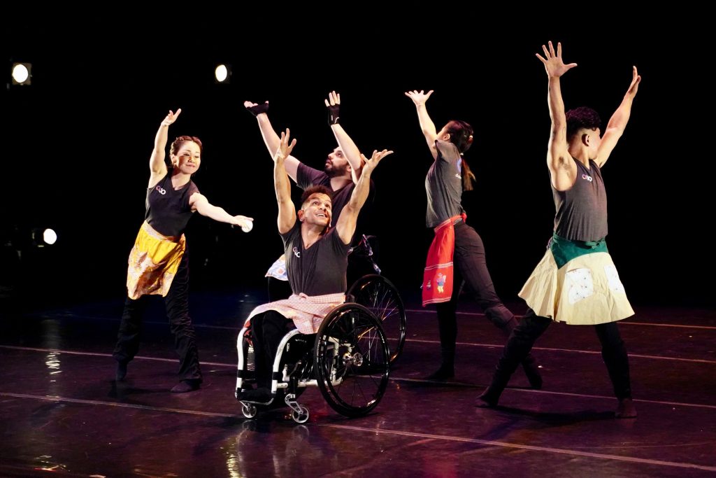 Five disabled and non-disabled dancers raise their arms up as they dance in a circle; they wear black AXIS t-shirts and multi-colored aprons.