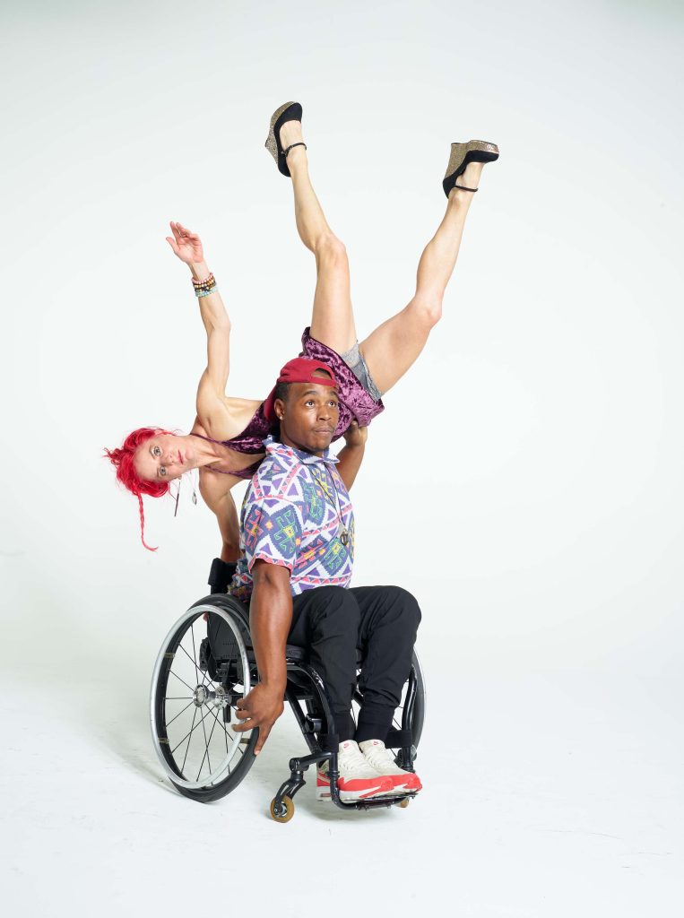 Sonsherée balances the side of her torso on DeMarco’s shoulder; she extends her upper and lower limbs upwards while DeMarco holds her and gazes upwards. Sonsherée has bright red hair and wears a purple velvet dress; DeMarco wears a patterned T-shirt, red cap and orange & white sneakers and sits in his manual wheelchair.