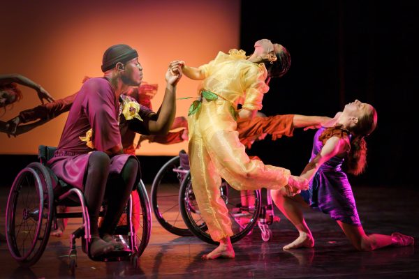 Disabled and Non-disabled dancers reach across space to hold each other's upper and lower limbs in a circular chain. Flower-inspired costumes are worn in bright hues of yellow, maroon, orange and purple.