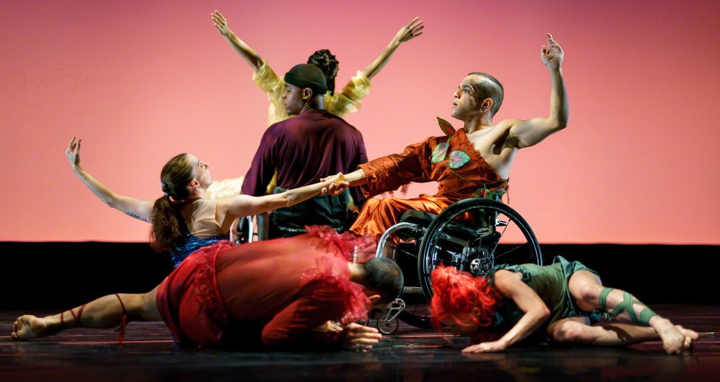 Six disabled and non-disabled dancers pose from low to high levels in symmetry and counterbalance to create a floral tableau. Dancers wear bright costumes with maroon, yellow, green and aquamarine coloring.