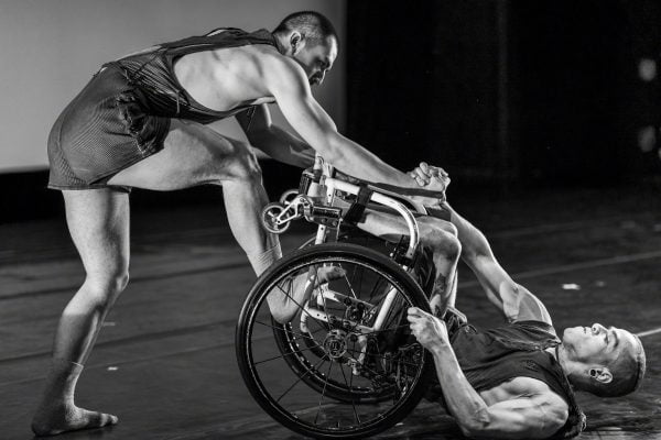 Janpistar lies on their back in their wheelchair, they grasp the hand of Erik Debono who is lifting them off the floor. Erik places one foot on Janpi's chair for stability. Both dancers have strong, defined muscles and wear black sleeveless tops and shorts.