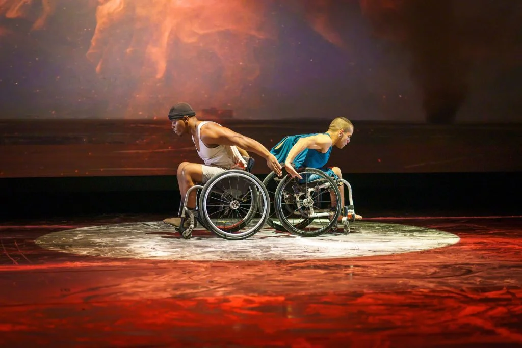 Janpi and DeMarco face back-to-back and begin to wheel away from each other in their wheelchairs. They are lit by a white circle of light on a dance floor textured by baking soda. The rest of the floor is lit red with fiery projections on the back wall.