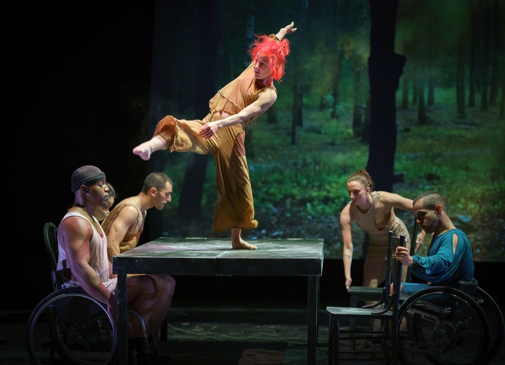 Sonsherée Giles dances on top of a large rectangular table; she lifts one leg forward and reaches one arm back in a diagonal. Four disabled and nondisabled dancers sit on the table to her left and right. Behind them is a projection of trees in a forest.