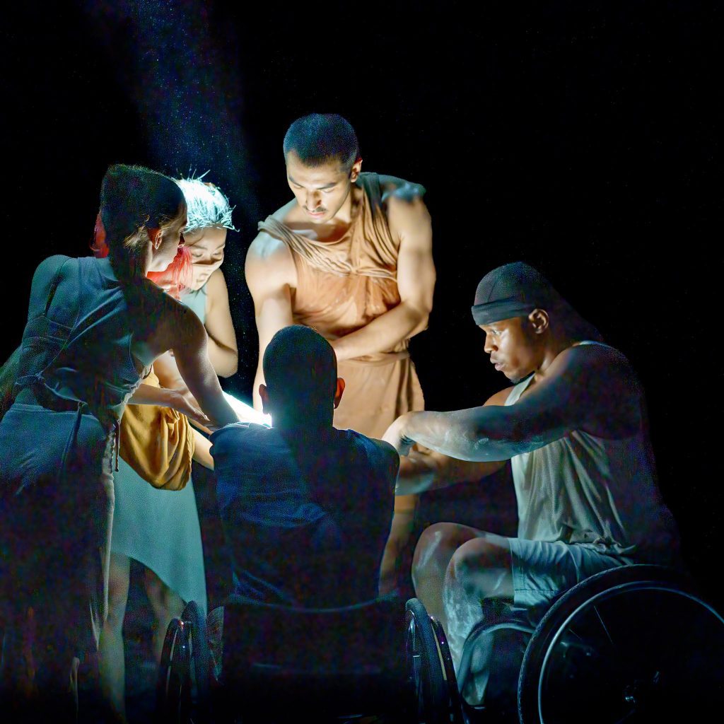 Six disabled and non-disabled dancers bring their upper limbs together on stage, as a concentrated beam of lights shines on their hands. The light captures fine white particles of baking soda in the air.