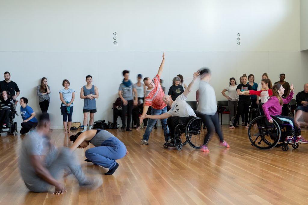 An artistically blurred image of disabled and non-disabled dancers partnering together in a workshop. Some dancers watch on the side while other dancers partner with each other on a wooden dance floor.